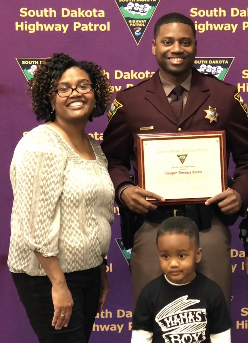Trooper Terrence Davis in uniform holding a South Dakota Highway Patrol award plaque, with his wife and young son